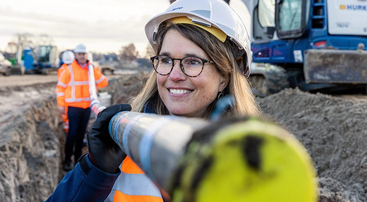 Smiling woman in workwear holding a pipe together with a man in the background (photo)